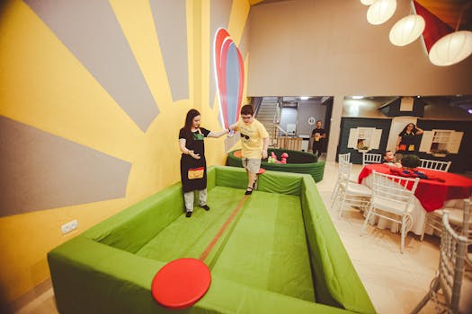 A child plays on a balance beam indoors, guided by an adult in a colorful playroom setting.
